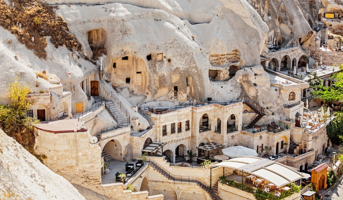 A traditional cave hotel in Cappadocia built into the soft volcanic cliffs, featuring arched stone architecture, terraces, and carved rock rooms. The unique design combines ancient cave dwellings with modern amenities, offering visitors an unforgettable stay.