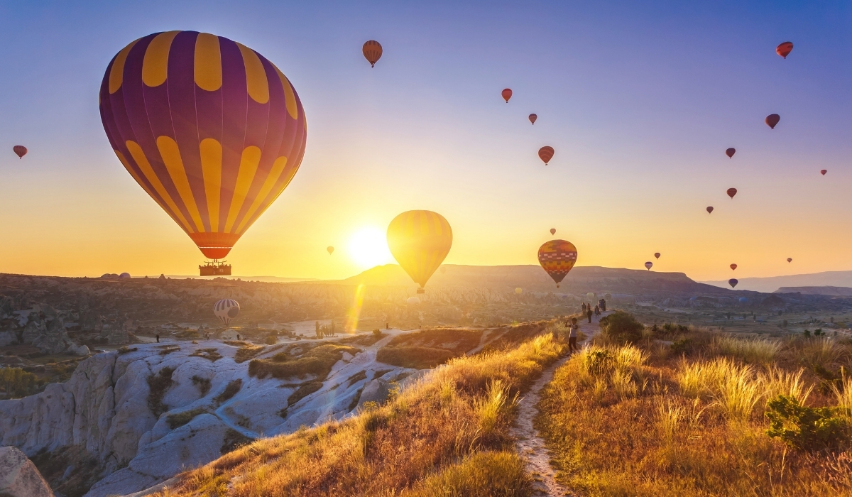 Multiple hot air balloons soar above the valleys of Cappadocia as the sun rises on the horizon. The warm golden sky and rugged terrain below make this one of the region’s most breathtaking scenes.