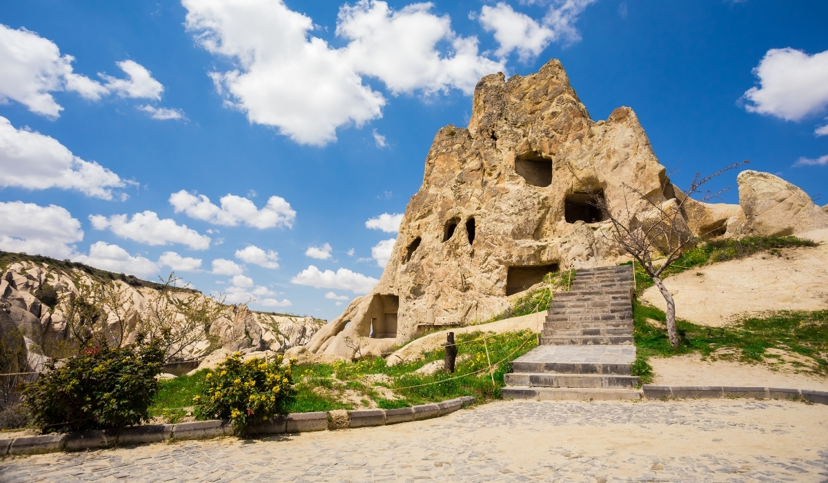 A historic rock-carved cave church in Cappadocia with multiple openings in the volcanic rock face. The stone steps leading up to the entrance highlight the impressive craftsmanship and cultural heritage of early Christian settlements in the region.