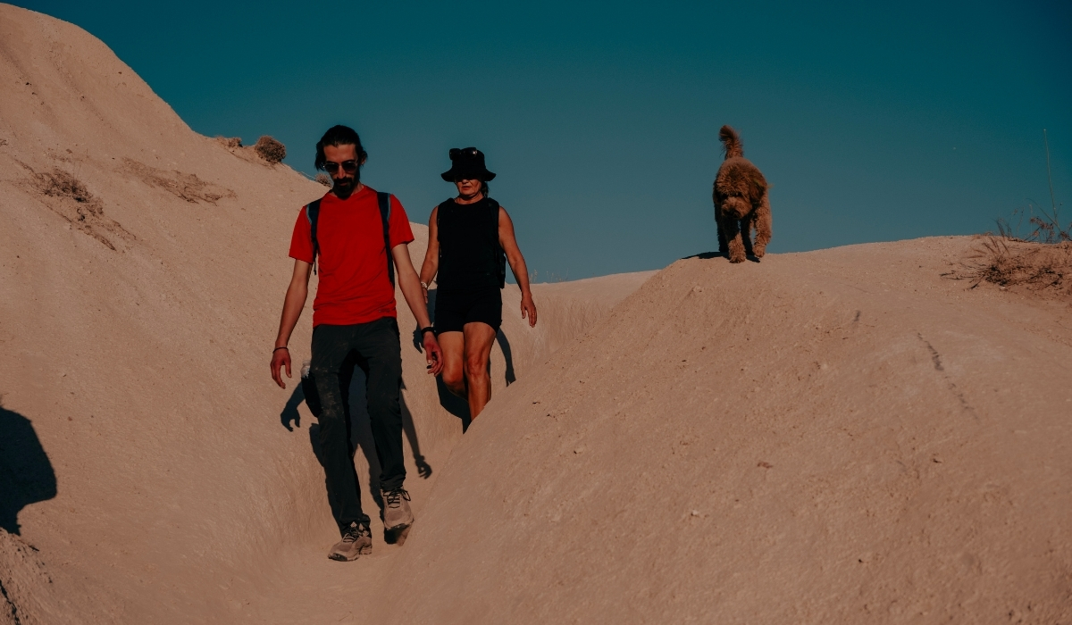 Two travelers hiking through Cappadocia’s rugged terrain with a dog, surrounded by volcanic rock formations under a clear blue sky. The scene highlights the adventurous hiking opportunities and natural beauty of Cappadocia’s valleys.